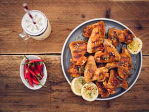Overhead shot of a plate of spicy chicken wings with a bowl of fresh red chillies and a summer beverage on a vintage wooden surface